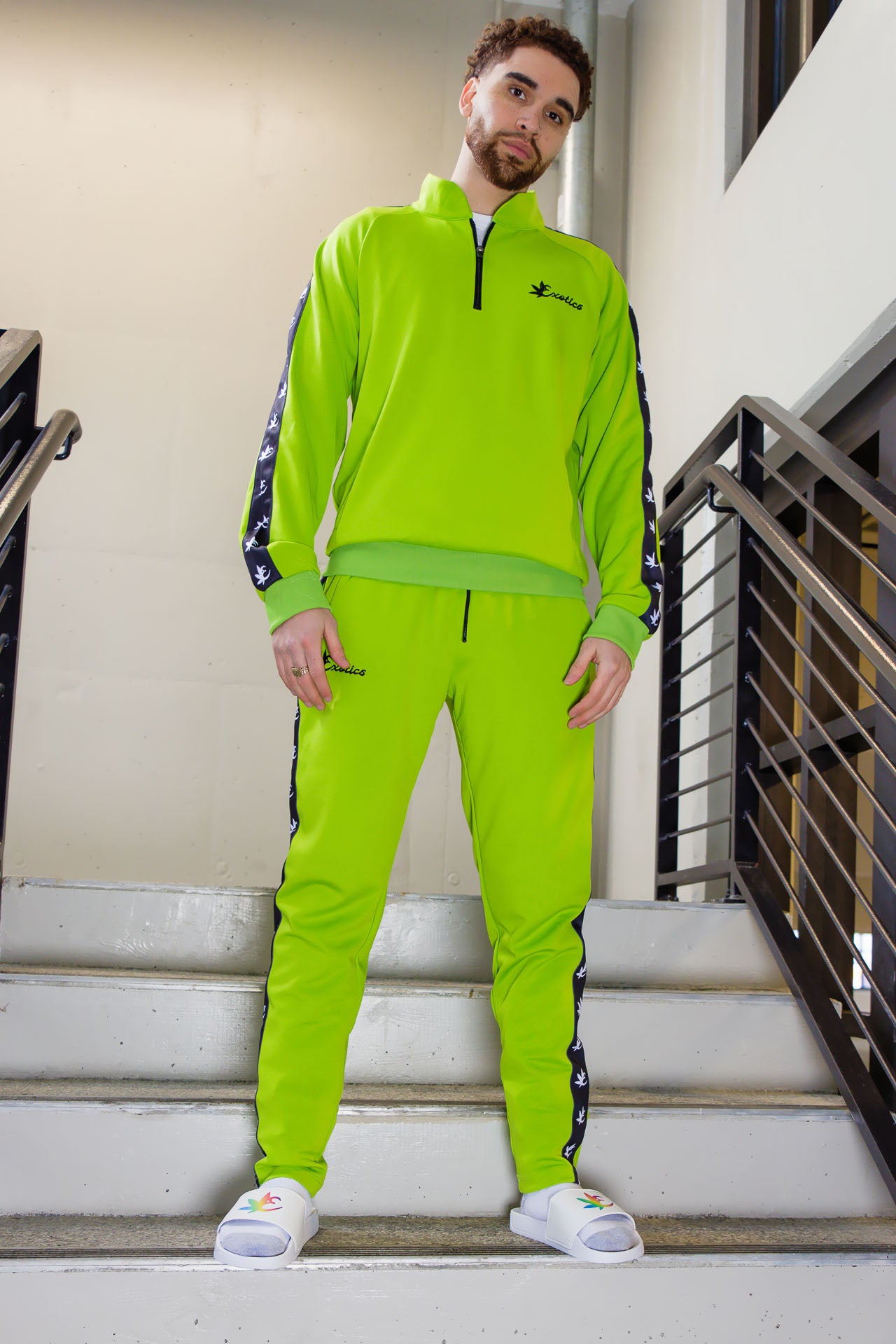 Man standing in stairwell wearing a green Exotics tracksuit and white slides